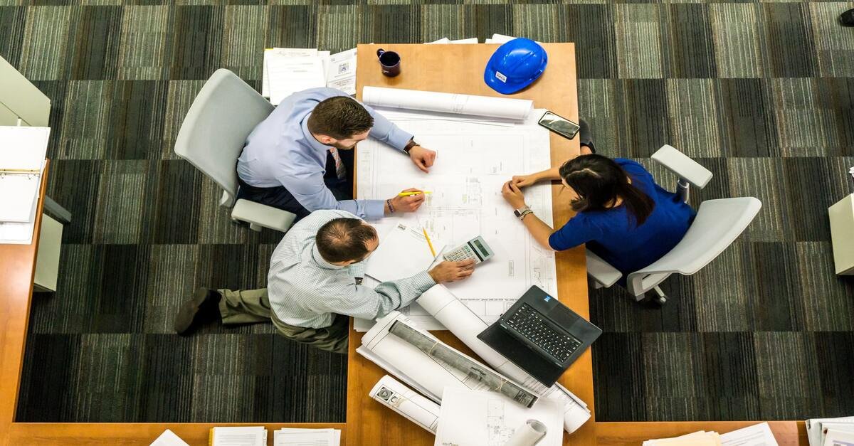 Birdseye view of two men and a women having a meeting with a laptop, calculator, large pieces of paper and a hard hat on the table