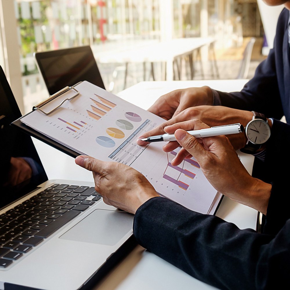 A picture of two men sat Infront of two laptops holding a clipboard with a piece of paper with different types of graphs on it