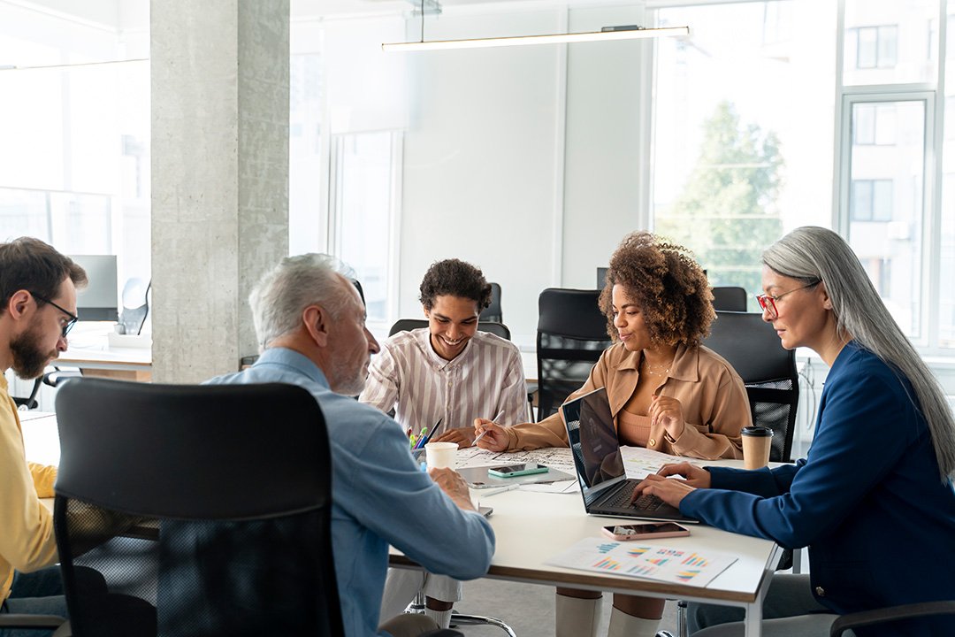 Relaxed business meeting between 5 people sitting at meeting table in naturally lit room